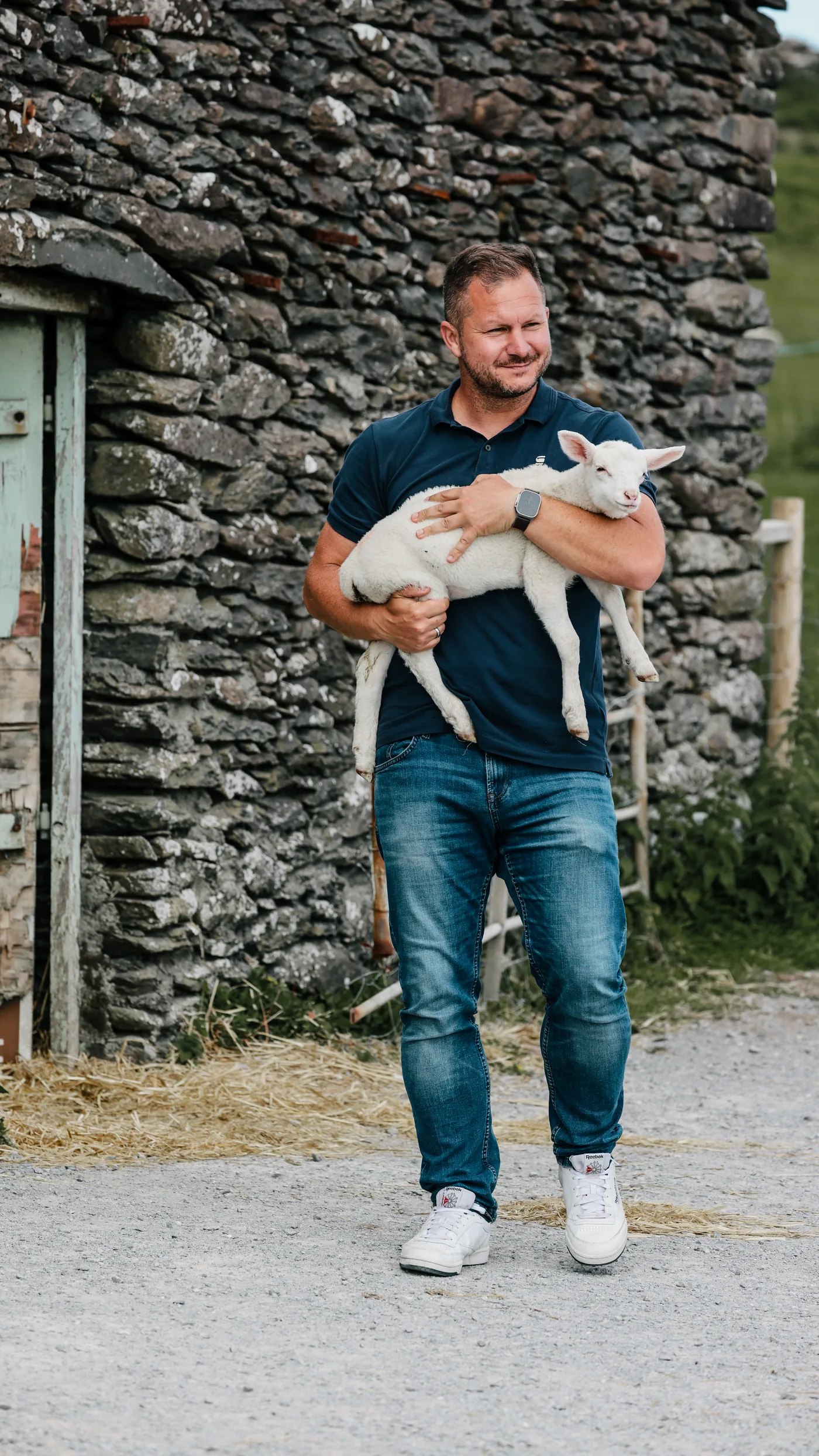 Benny mit einem Lamm auf dem Arm vor einer Steinmauer